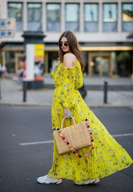 Модный лук: желтое платье-сарафан в пол с синими цветами, большие белые кроссовки, большая соломенная сумка с разноцветными помпонами и темные солнцезащитные очки