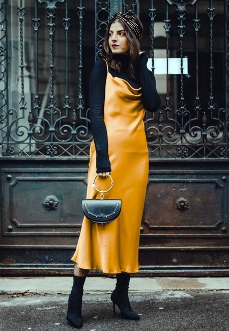 Модный лук: желтое атласное платье-комбинация длины миди, черная водолазка, черные замшевые сапоги на шпильке, маленькая черная кожаная сумочка полукруглой формы с золотой ручкой и цветная повязка