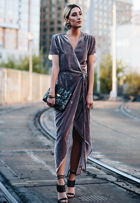 Модный лук: Серое бархатное платье-маллет на запах и черные босоножки на шпильке с завязками, блестящая сумка-конверт, большие круглые серьги, металлические браслеты 